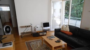 Un salon avec un canapé noir, une table basse en bois, une imprimante, un ordinateur et une grande fenêtre.