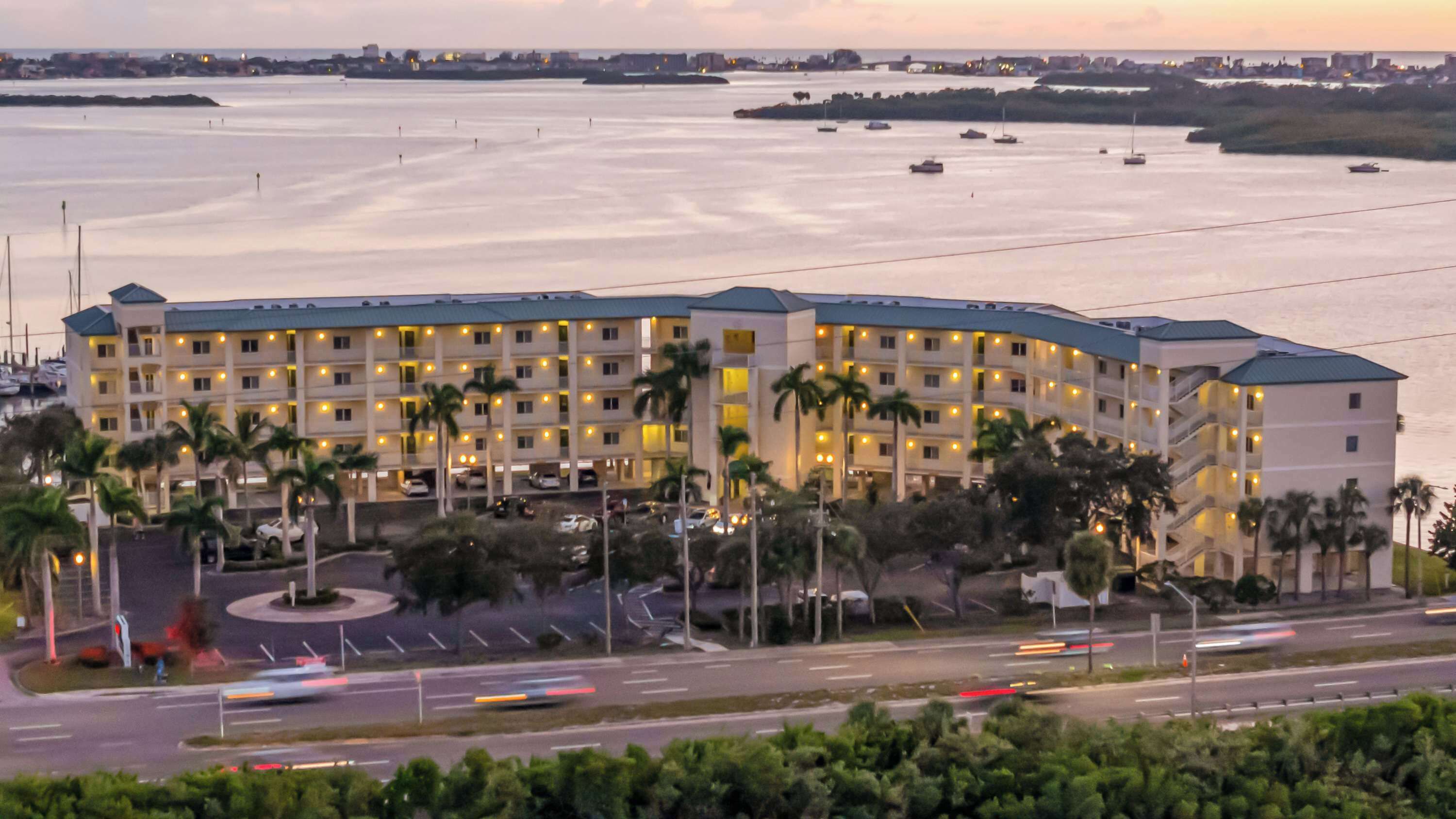 Un hotel de varios pisos con vista al agua, que cuenta con un estacionamiento y palmeras en primer plano.