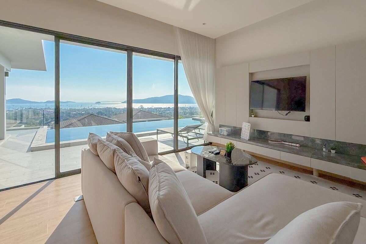A modern living room with a large sofa, a TV mounted on the wall, and a view of a pool and mountains.