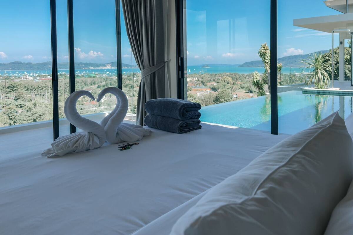 A hotel room with a large bed, a heart-shaped towel arrangement, a view of the sea, and a pool.