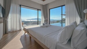 Quarto moderno com uma cama grande, duas janelas com cortinas e vista para a cidade e montanhas.