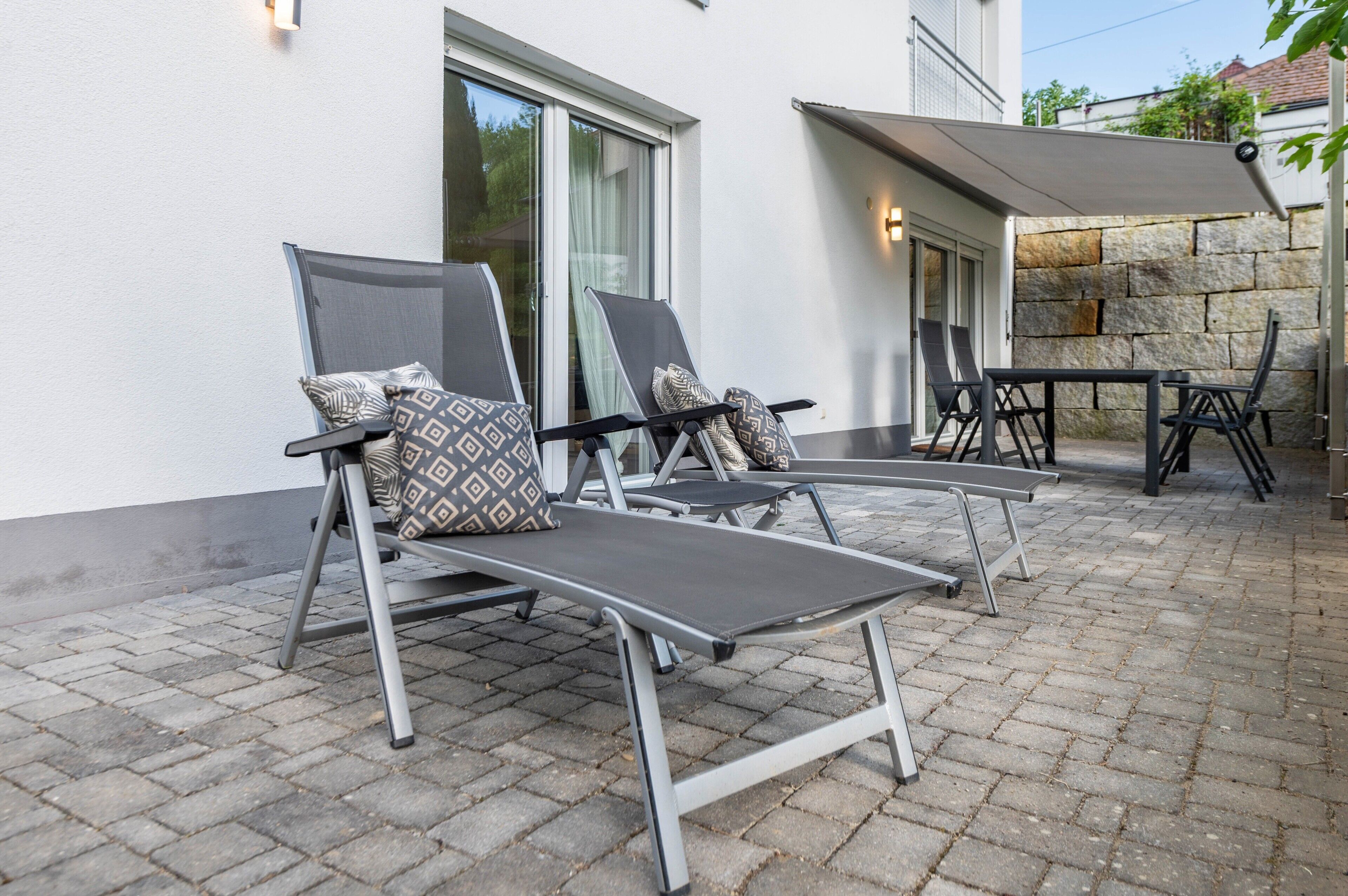 Eine Terrasse mit zwei Lounge-Stühlen und einem Tisch, einer Steinmauer und einem überdachten Bereich mit einer Lampe.