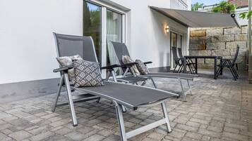 Eine Terrasse mit zwei Lounge-Stühlen und einem Tisch, einer Steinmauer und einem überdachten Bereich mit einer Lampe.