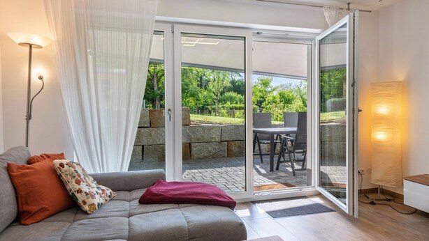 En moderne stue med sofa, bord og en skydedør, der fører til en udendørs terrasse.