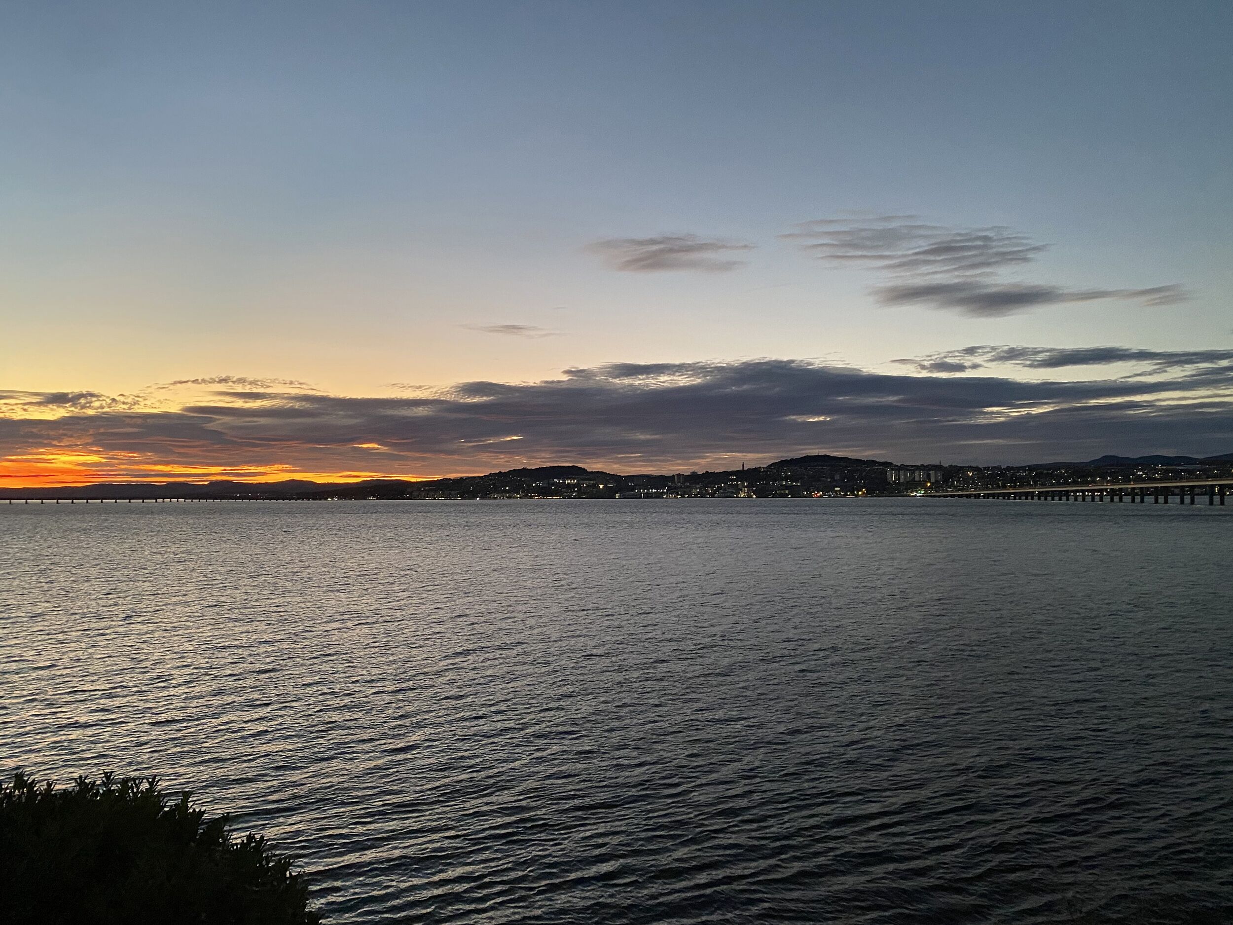 A sunset over a calm body of water with a bridge and distant cityscape.