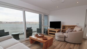 Una sala de estar moderna con un amplio ventanal, un televisor de pantalla plana, una mesa de centro de madera, un sofá color beige y un mueble de madera.