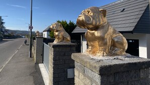 Una calle con dos estatuas doradas de bulldogs sobre un pedestal de piedra, una señal de límite de velocidad y casas al fondo.