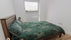 Un dormitorio con una cama de madera, una colcha estampada verde, vistas a una masa de agua a través de una ventana y una mesita de noche de madera con una lámpara.