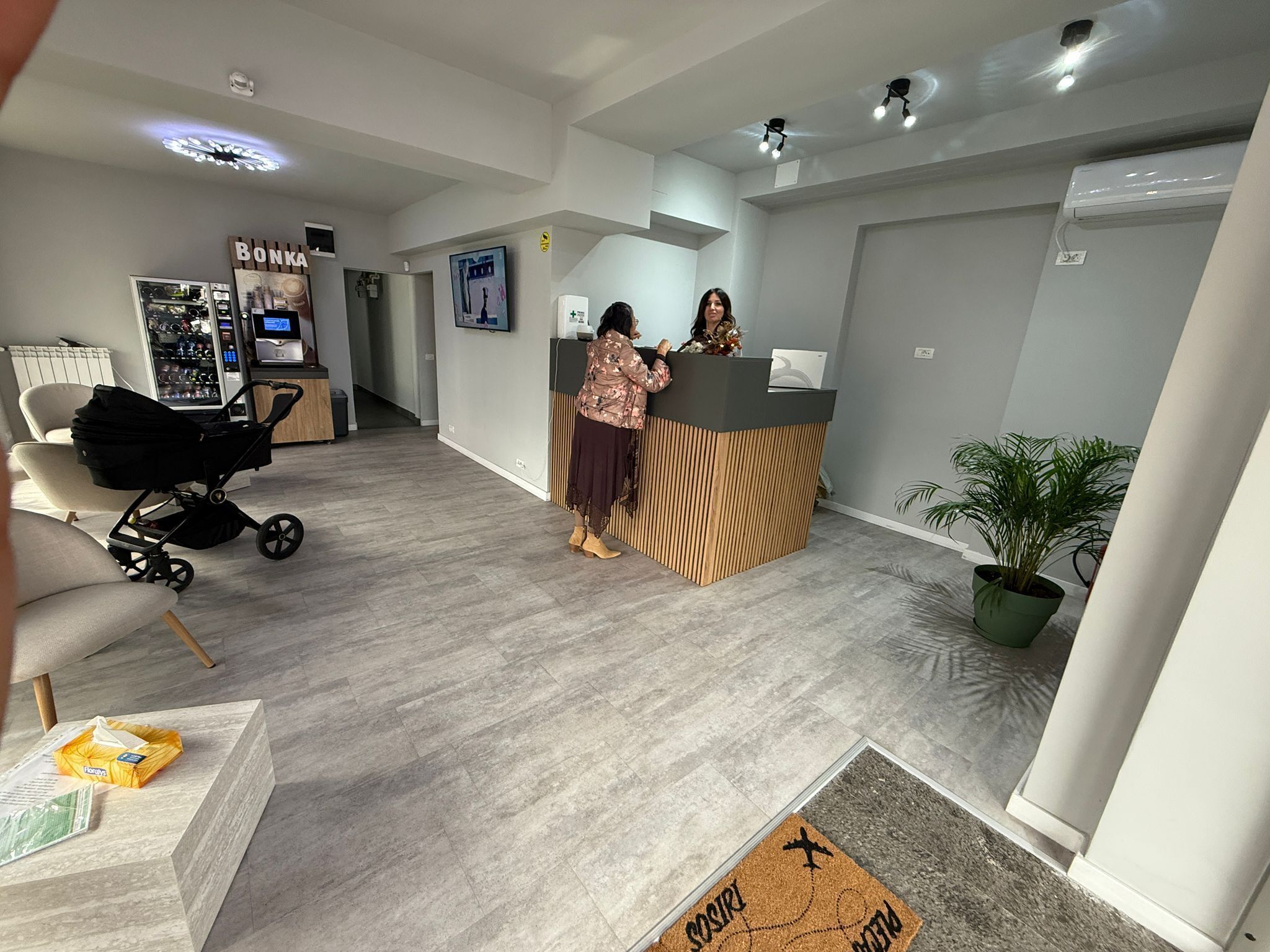 A reception area with a wooden reception desk, a refrigerator with beverages, a chair, a stroller, and a potted plant.