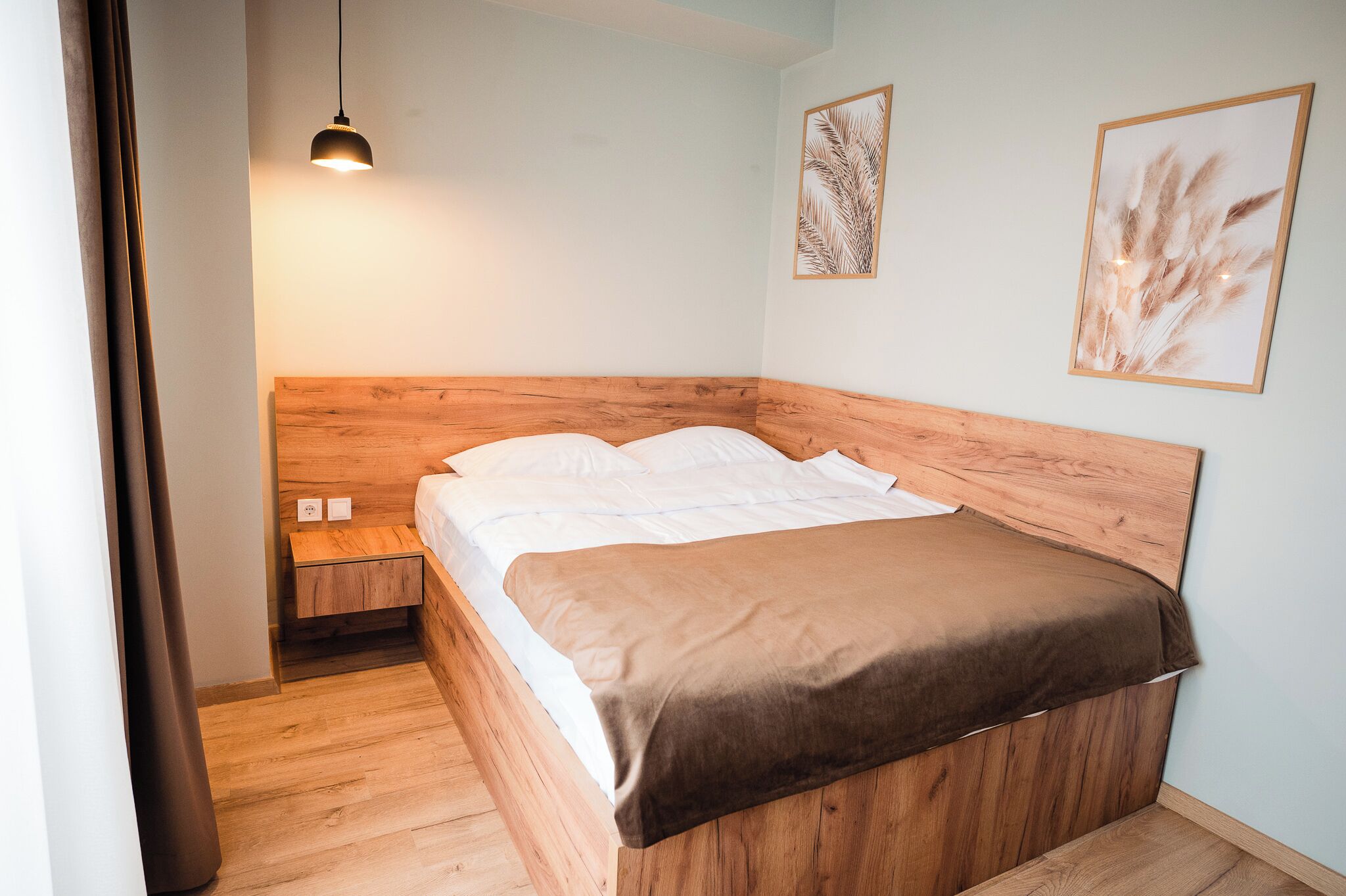 Un dormitorio con cabecera de madera, cama individual con ropa blanca, manta marrón, mesita de noche con lámpara y dos cuadros enmarcados.