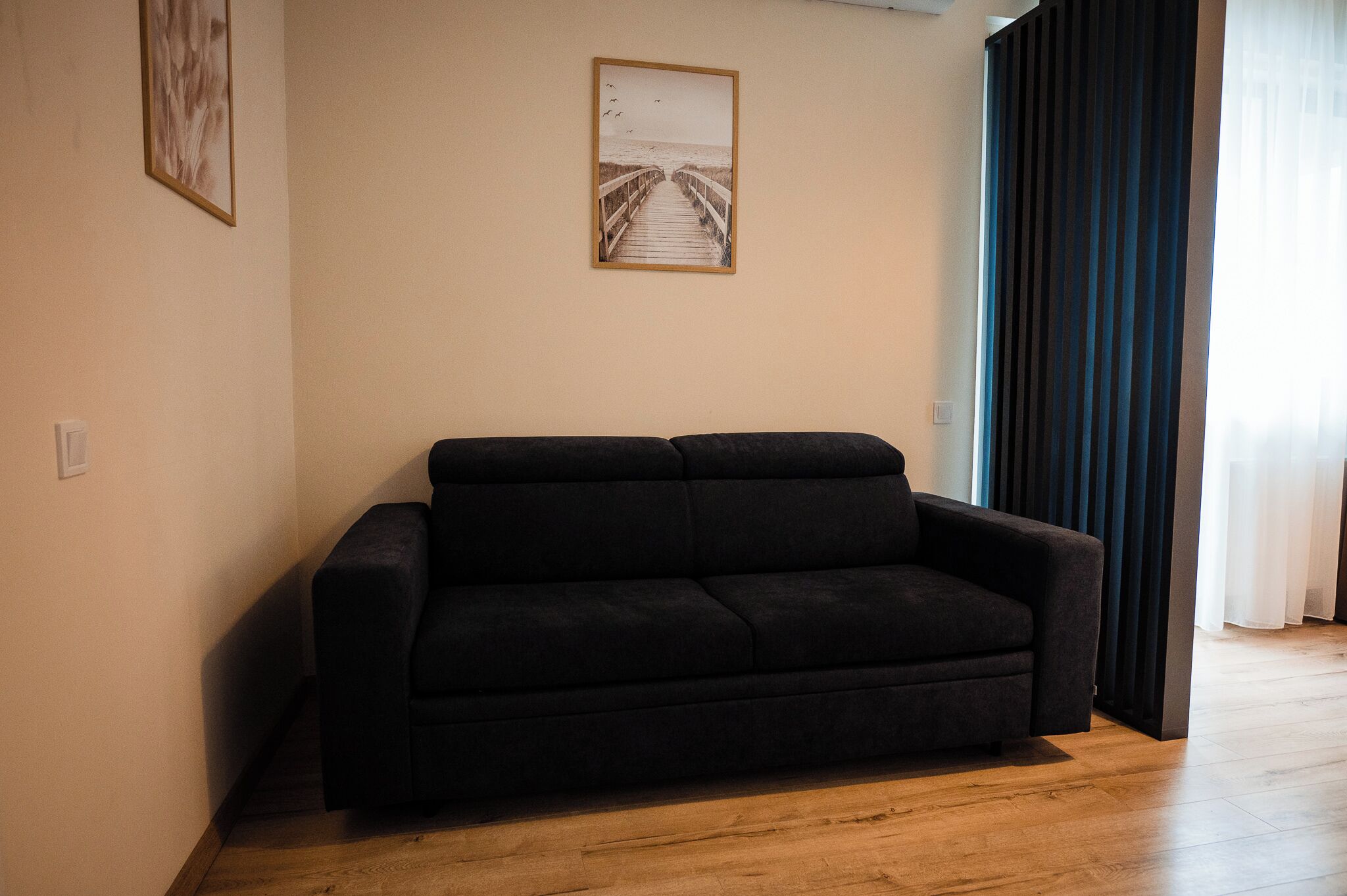A dark-colored sofa in a room with wooden flooring, a framed picture on the wall, and a window with dark curtains.