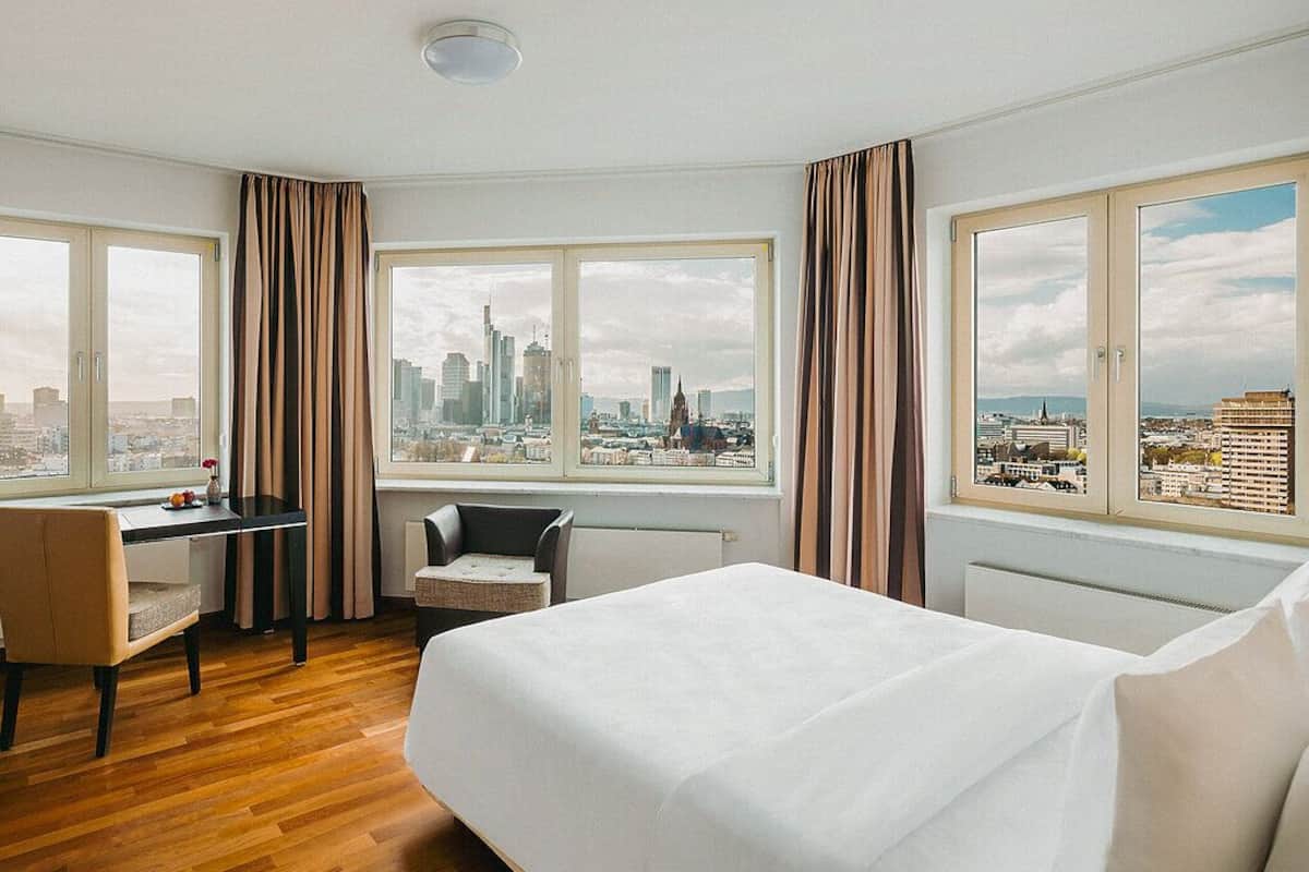 Een hotelkamer met een bed, een bureau, een stoel en grote ramen met uitzicht over de stad.
