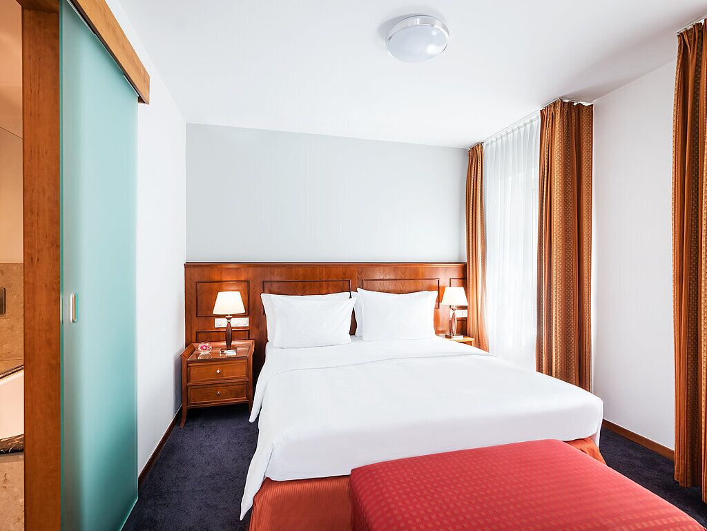 A hotel room with a large bed, wooden headboard, two bedside tables with lamps, and a window with curtains.