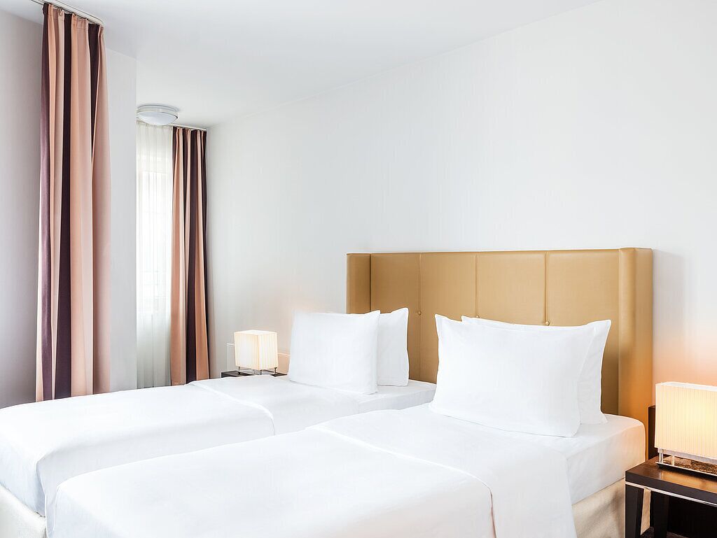 Hotellihuone, jossa on kaksi sänkyä, sängynpääty, yöpöydät lampuineen ja ikkuna, jossa on verhot.