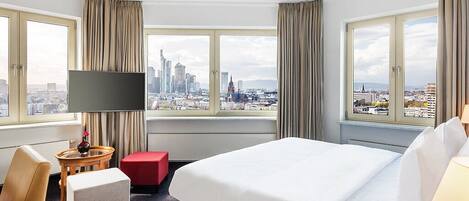 Habitación de hotel con una cama grande, televisión, una mesita, un reposapiés rojo y vistas a un horizonte urbano.