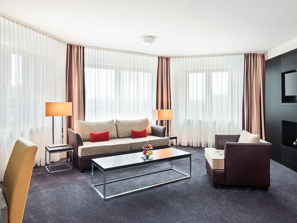 A modern hotel room with a sofa, armchairs, a coffee table, and a TV.