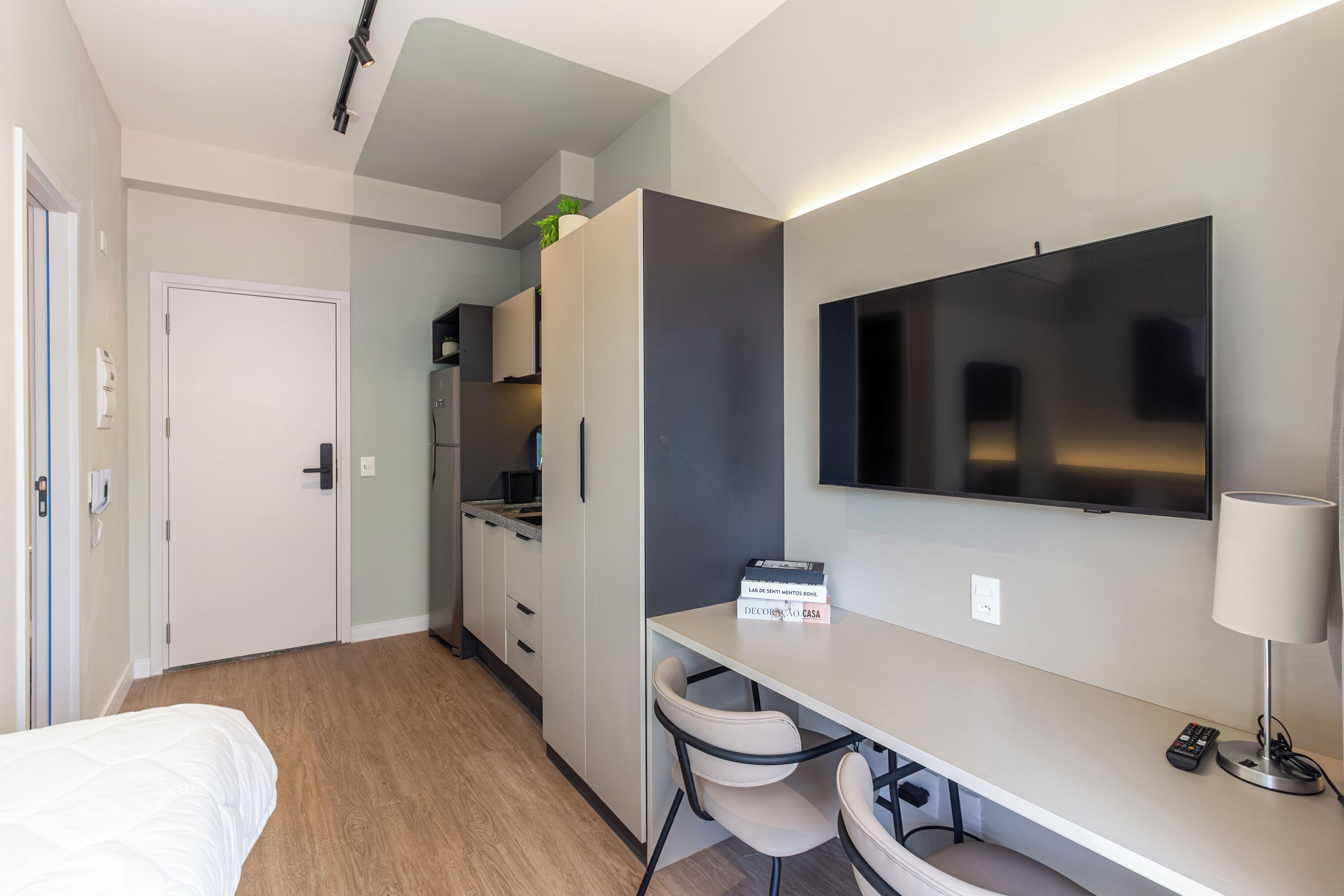 A hotel room with a bed, a desk with a chair, a TV, and a lamp. There is a door and a kitchenette in the background.