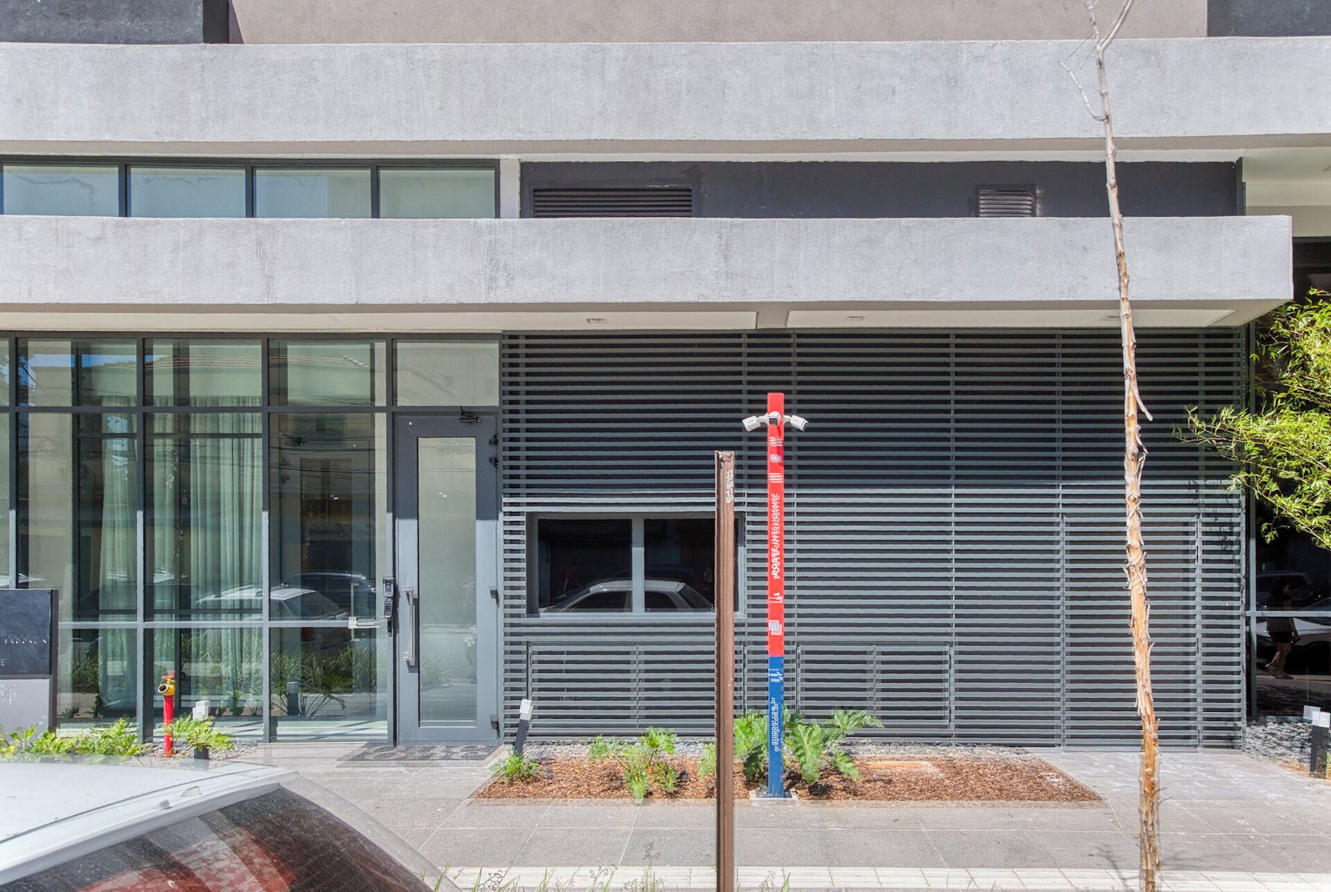 Un bâtiment moderne doté d’une entrée en verre, d’une grille métallique et d’un petit jardin agrémenté de plantes.