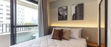 Habitación de hotel con una cama grande, una mesita pequeña y vistas a edificios desde un balcón.