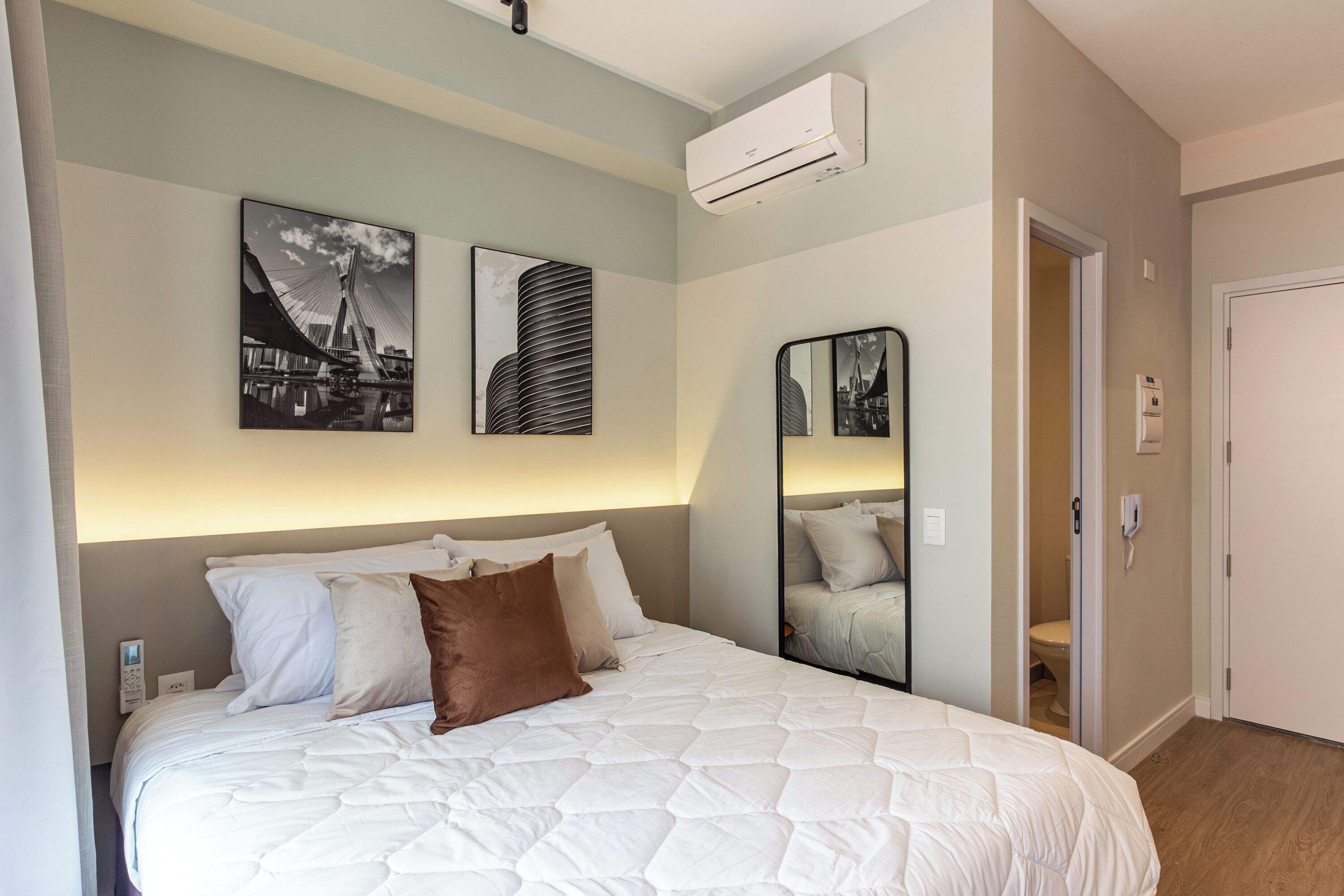 Una cama bien hecha con un edredón blanco acolchado, una almohada marrón y un aire acondicionado fijado a la pared. Hay dos cuadros en la pared, uno de un puente y otro de un edificio. También hay un espejo montado en la pared.
