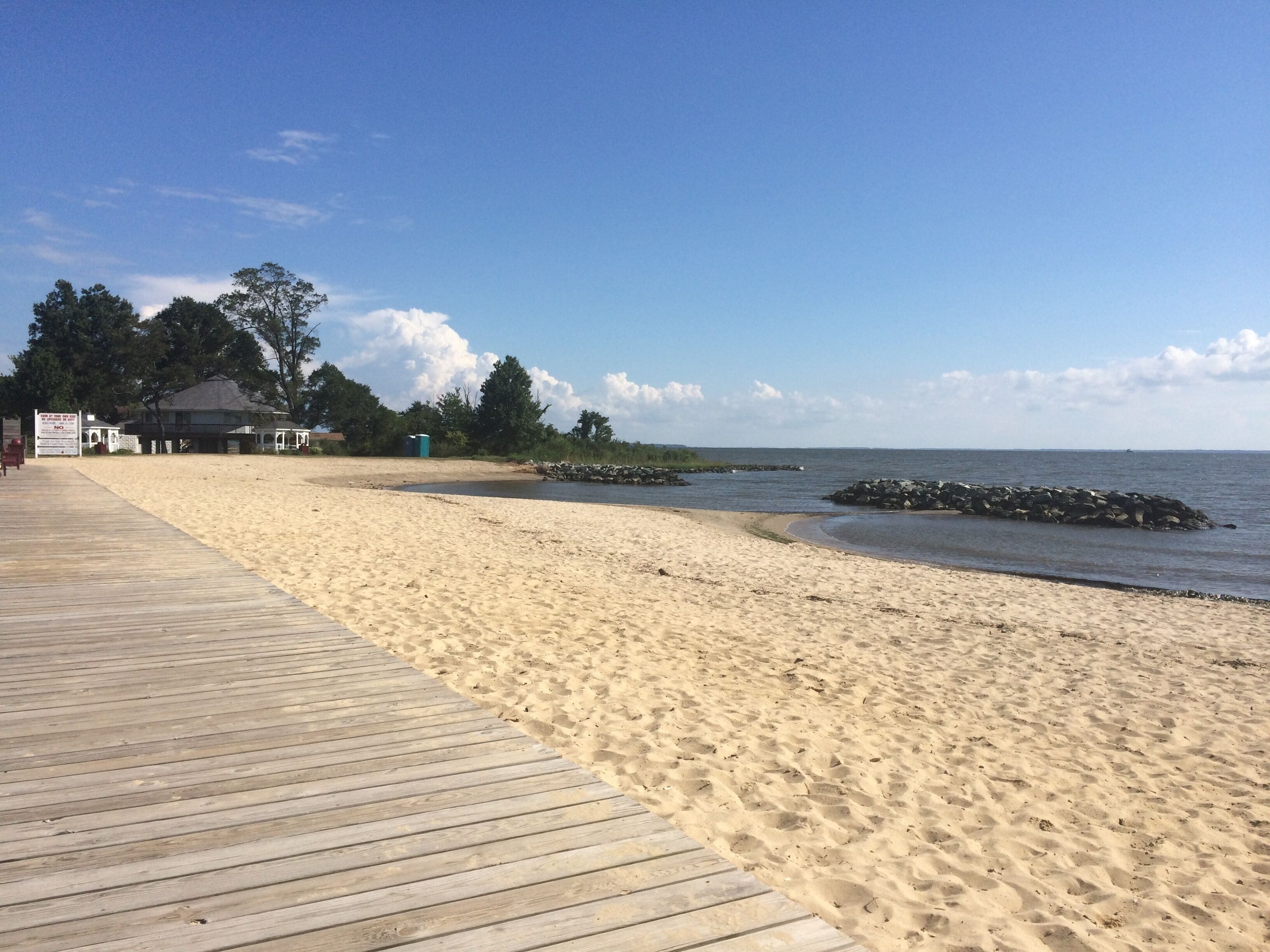 Ein hölzerner Steg führt zu einem Sandstrand, mit einem ruhigen Meer, einem Felsen und vereinzelten Bäumen im Hintergrund.