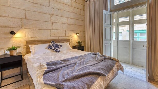 Quarto com parede de pedra, cama com travesseiro estampado, duas mesas de cabeceira com abajures e vista para o exterior através de uma porta.
