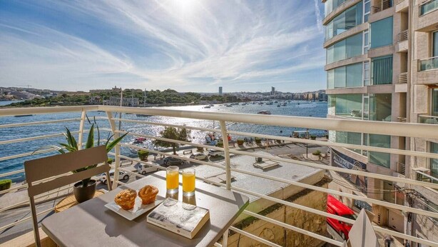 Een balkon met een ontbijttafel, met uitzicht op een jachthaven met boten en een stadsgezicht.