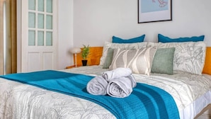 Een net opgemaakt bed met een blauwe deken en opgerolde handdoeken, een kamerplant en een lijst met een kunstwerk aan de muur.