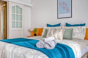 Una cama bien hecha con una manta azul y toallas enrolladas, una planta en maceta y una obra de arte enmarcada en la pared.