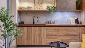 Una cucina moderna con mobili in legno, un lavello in acciaio inox e una macchina da caffè. Due lampade a sospensione, una stampa botanica incorniciata e una piccola pianta sul piano di lavoro.