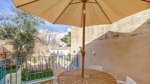 Un tavolo rotondo in legno con un ombrellone color beige, sedie bianche e una ringhiera blu in un cortile con un muro di pietra e vegetazione.