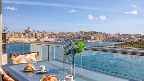 Een balkon met een glazen tafel, twee borden gebak, een kop koffie en een bloevas. Op de achtergrond een stadsgezicht met historische gebouwen en een wateroppervlak.