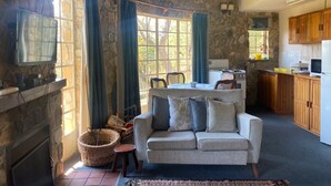 A cozy living room with a stone wall, a fireplace, a sofa, and a kitchen area with wooden cabinets and a microwave. - Beautiful Round Shaped Cottage - 2094 (Juliasdale)