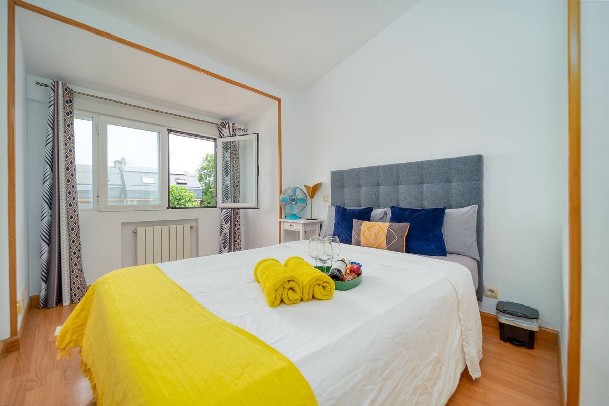 A hotel room with a large bed, a window with curtains, a bedside table with a lamp, and a small trash bin.
