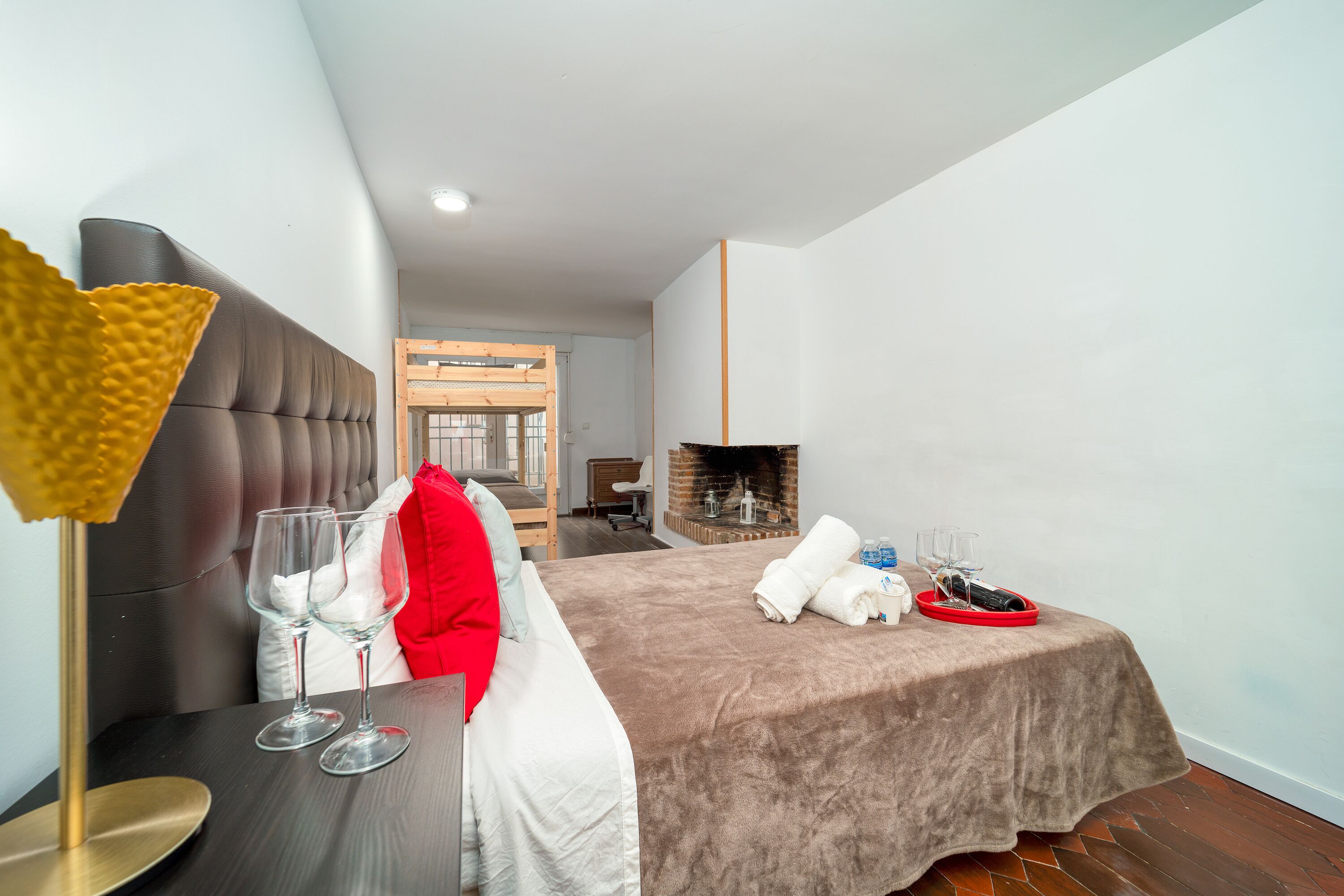 A modern hotel room with a large bed, a bedside table with wine glasses, a red pillow, and a fireplace.