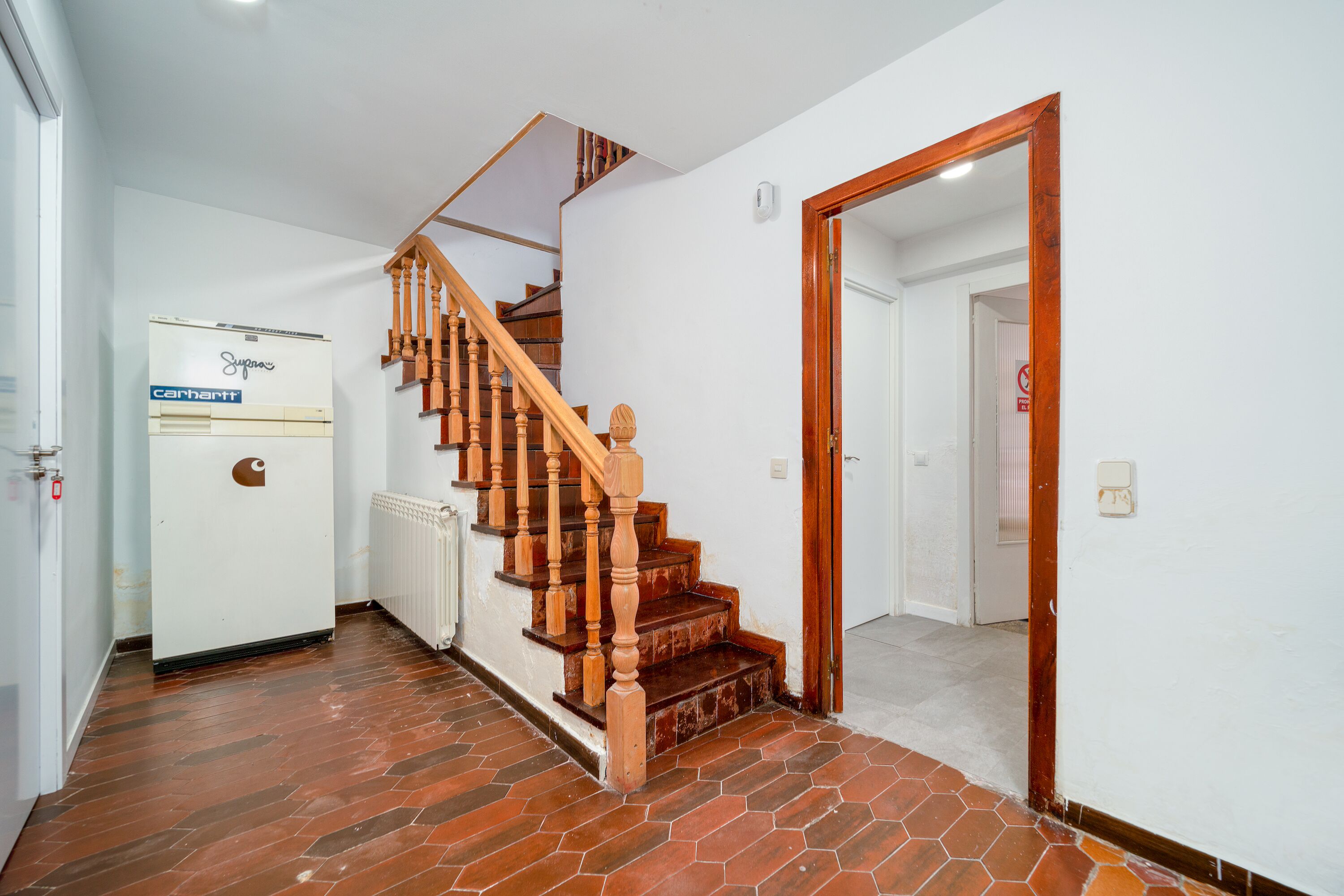 Una escalera con balaustres de madera que sube a un segundo piso, un radiador y una puerta con un cartel.