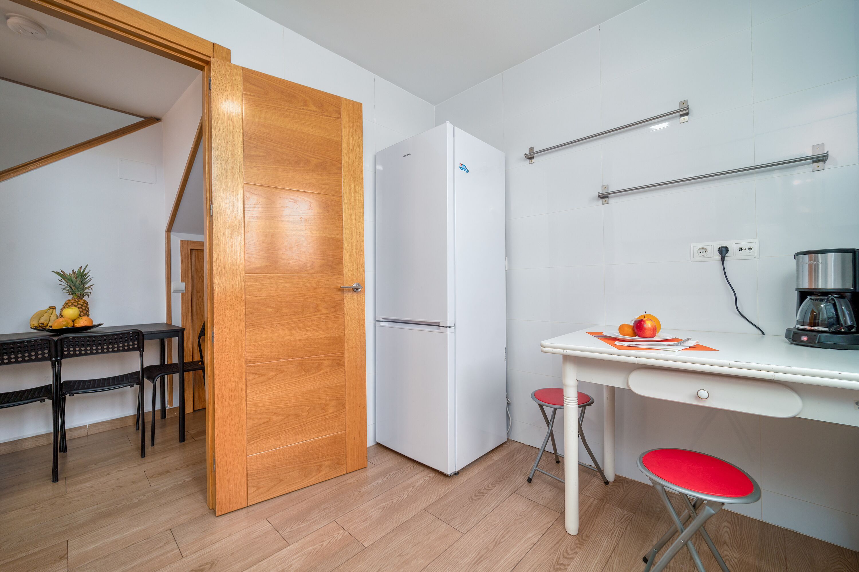 Uma cozinha compacta com porta de madeira, geladeira branca, mesa pequena com cadeiras e bancada branca com chaleira.