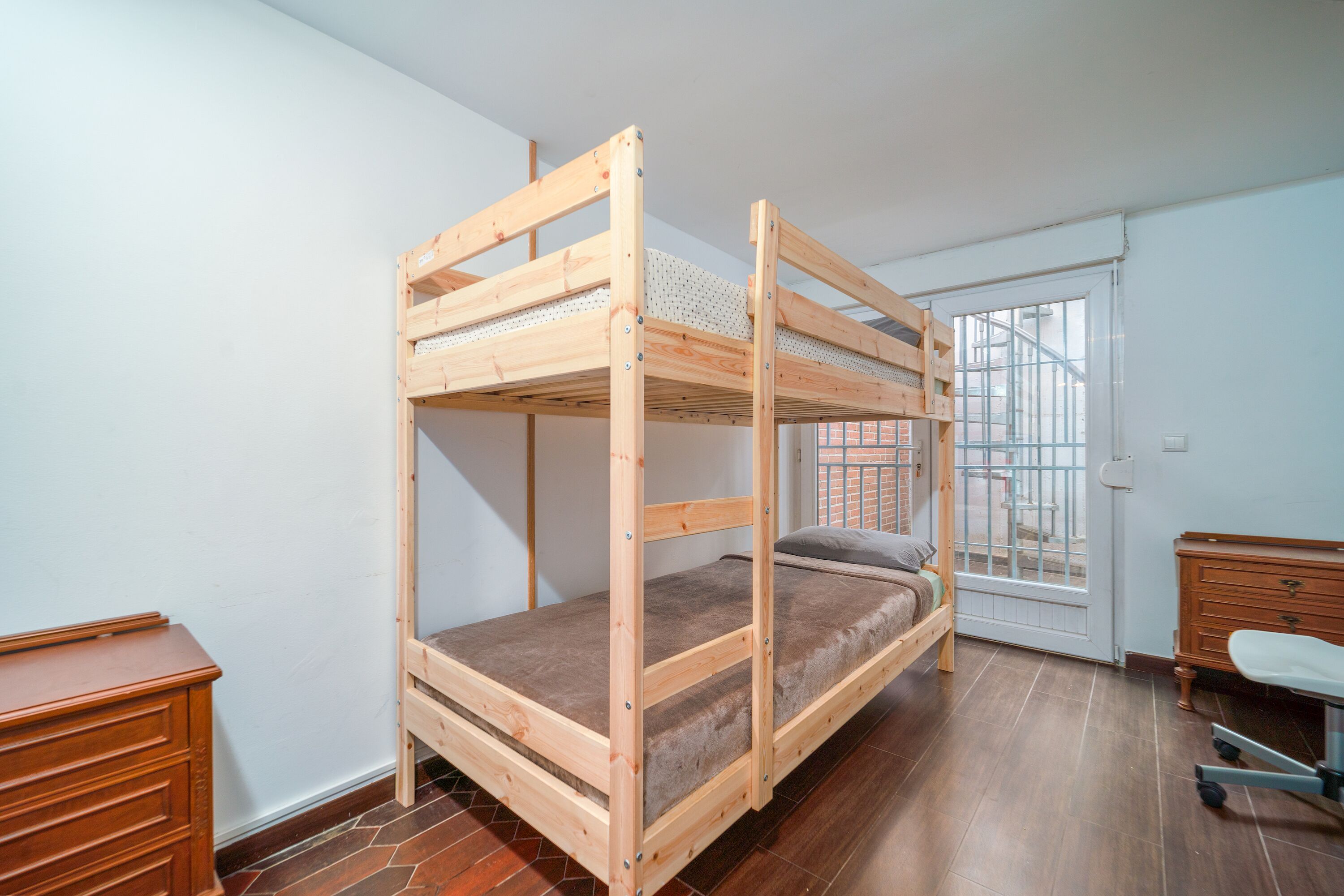 A bunk bed in a room with a wooden dresser, a desk, and a window with a view of a brick building.