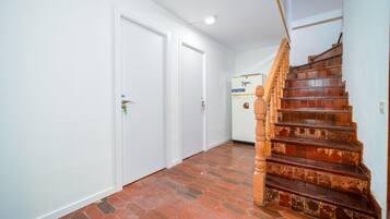 Un pasillo con paredes blancas, una escalera de madera junto a un muro de ladrillo y una puerta con cerradura.