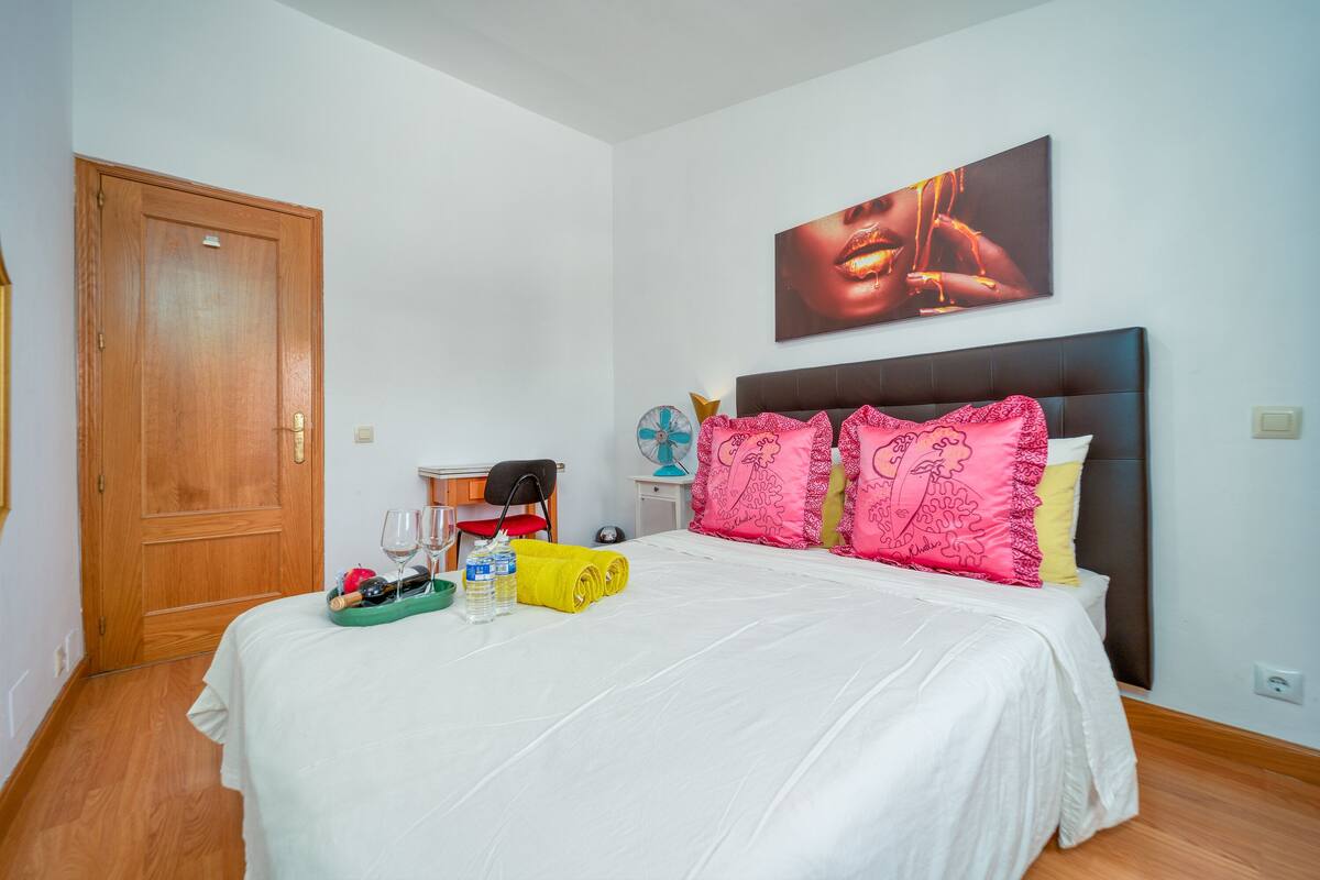 A neatly made bed with a white bedspread, a wooden door, and a wall-mounted artwork.