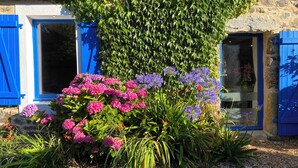 A building with blue shutters, a stone wall, and a garden with pink and purple flowers.