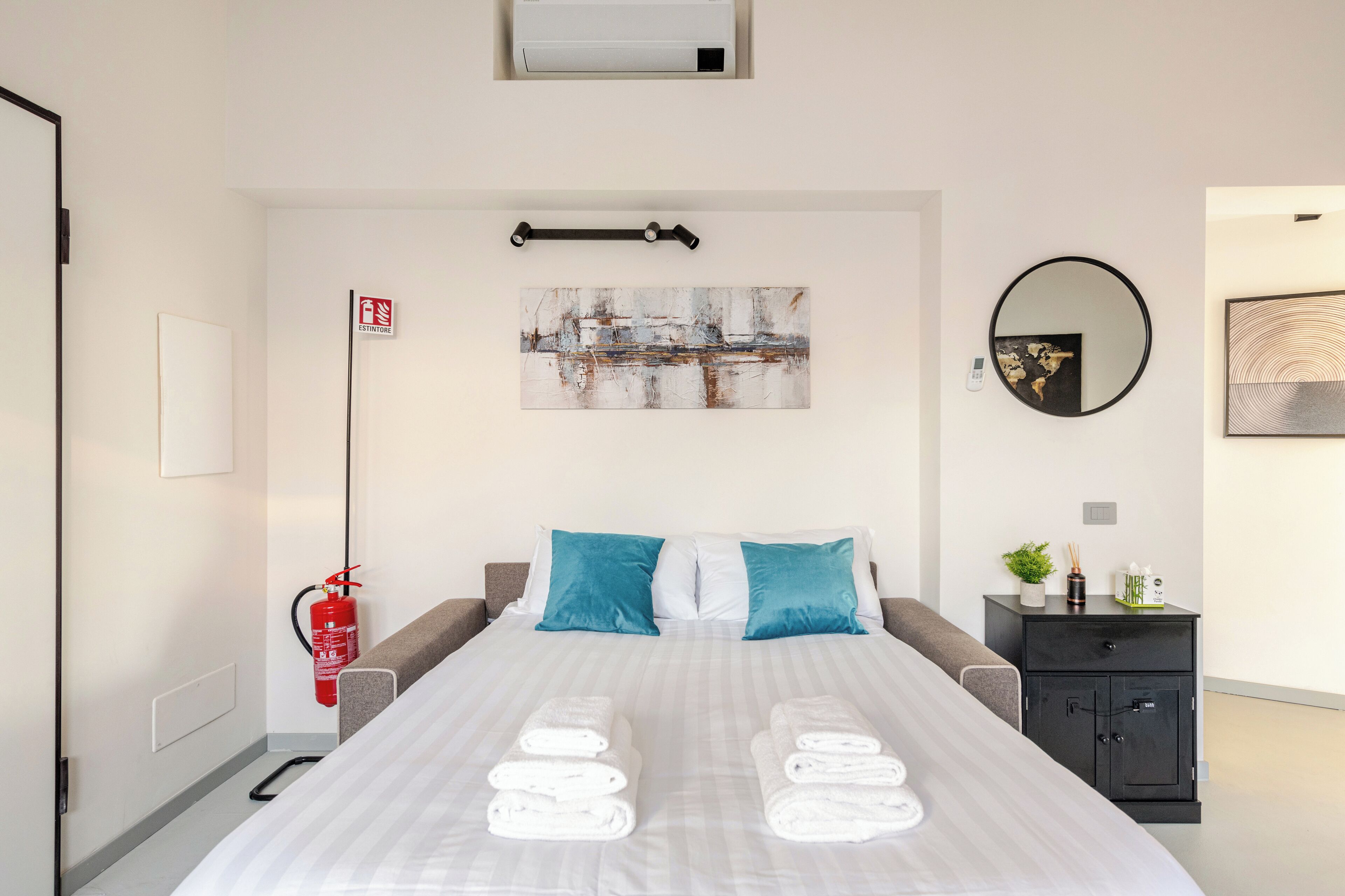 Quarto de hotel com cama, mesa de cabeceira, extintor de incêndio, quadro e espelho.