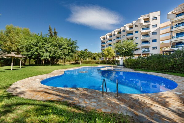 Piscina com azulejos de pedra, cercada por vegetação e um edifício de vários andares.