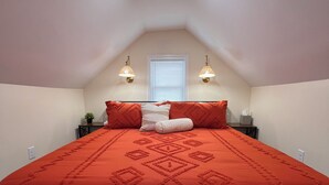 Un dormitorio con colcha roja, dos lámparas de noche, una ventana con persianas y una lámpara de techo.