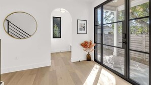 Ein geräumiges Zimmer mit Holzboden, einem großen Fenster mit Blick ins Freie, einem runden Spiegel an der Wand und einer Deckenleuchte.