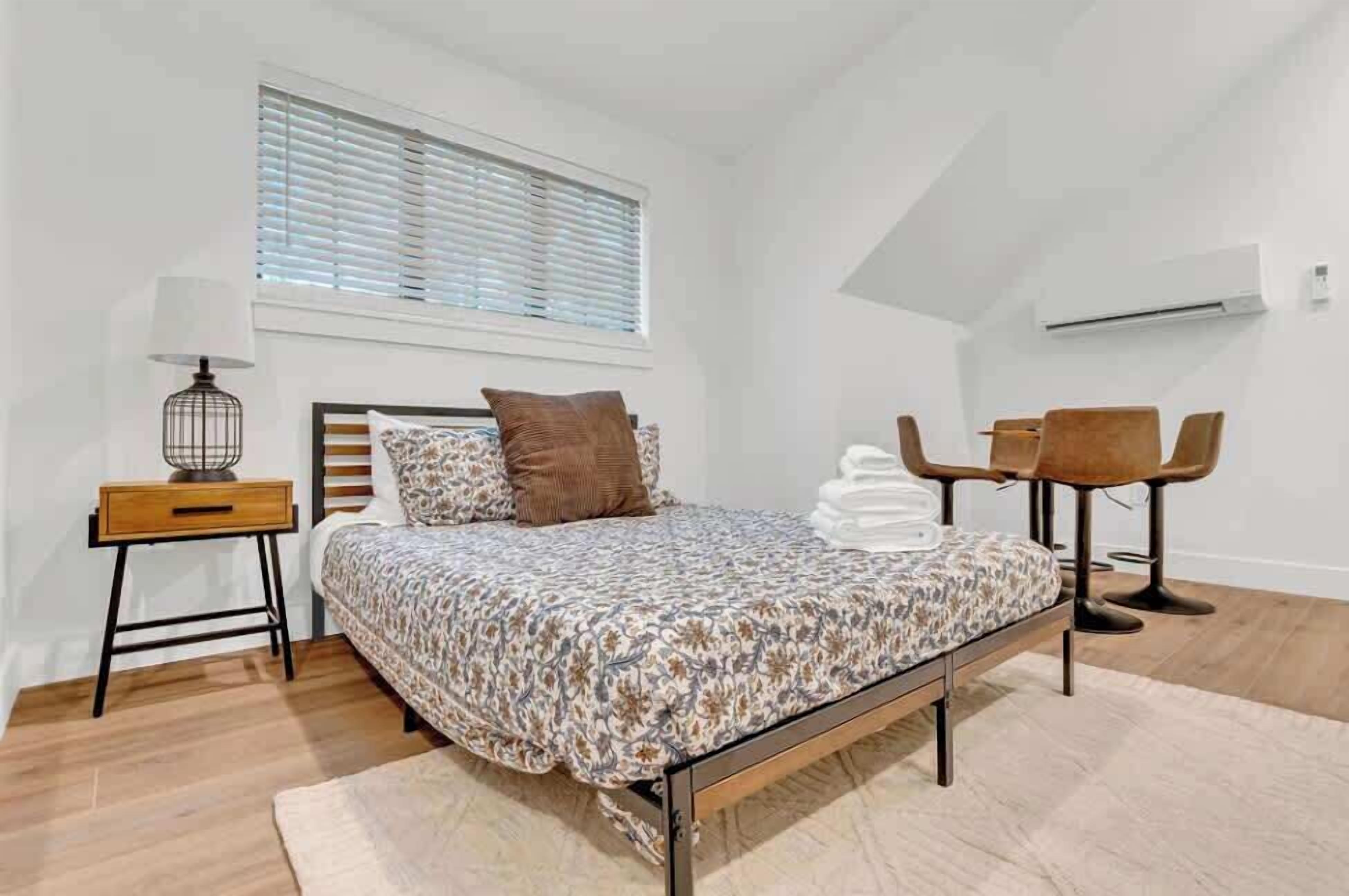 A bedroom with a bed, a nightstand, a lamp, a window with blinds, a chair, and an air conditioning unit.
