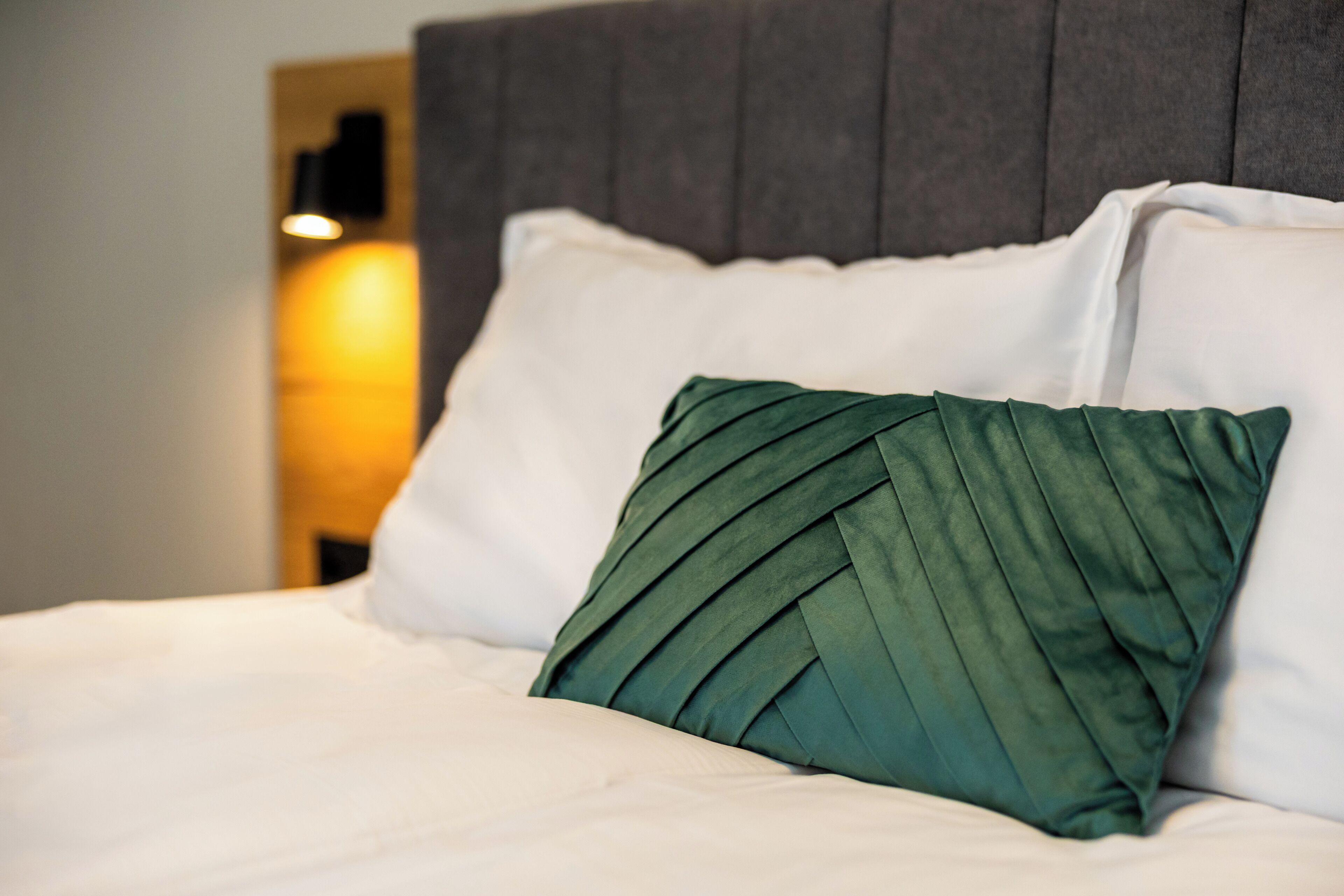 A bed with white bedding and a green pillow, a grey headboard, and a wall-mounted light fixture.