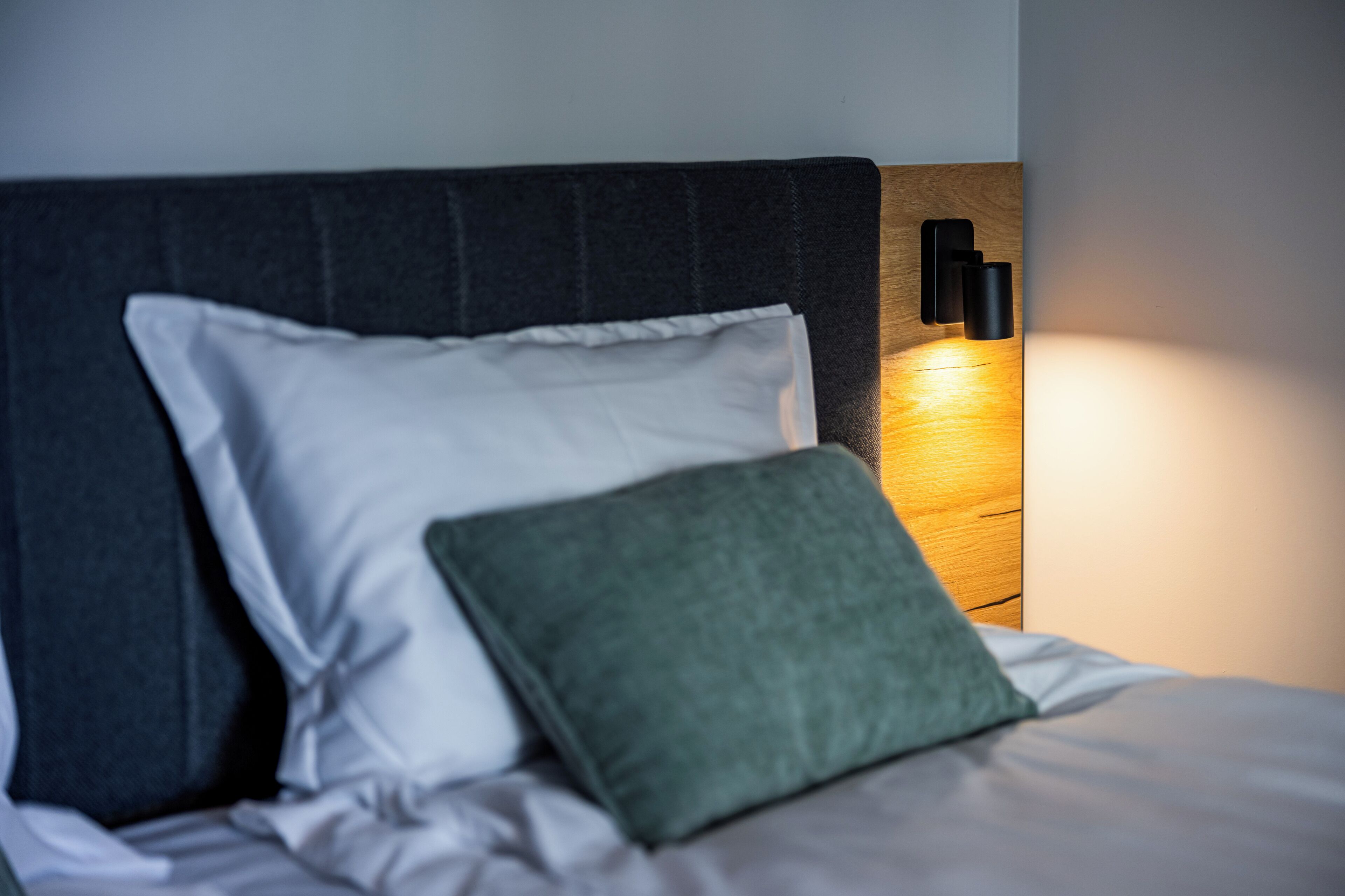 Una cama con almohadas blancas y verdes, un cabecero oscuro y una lámpara fijada a la pared.