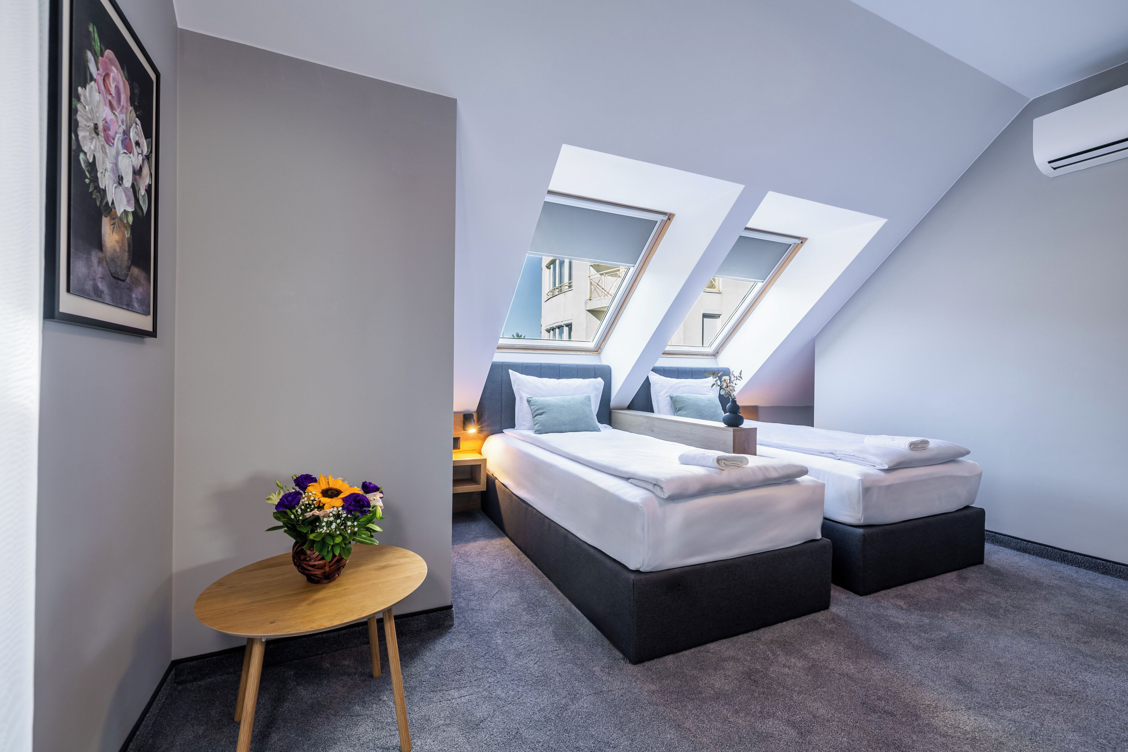A modern hotel room with a skylight, a bed, a small table with a vase of flowers, and a framed picture on the wall.