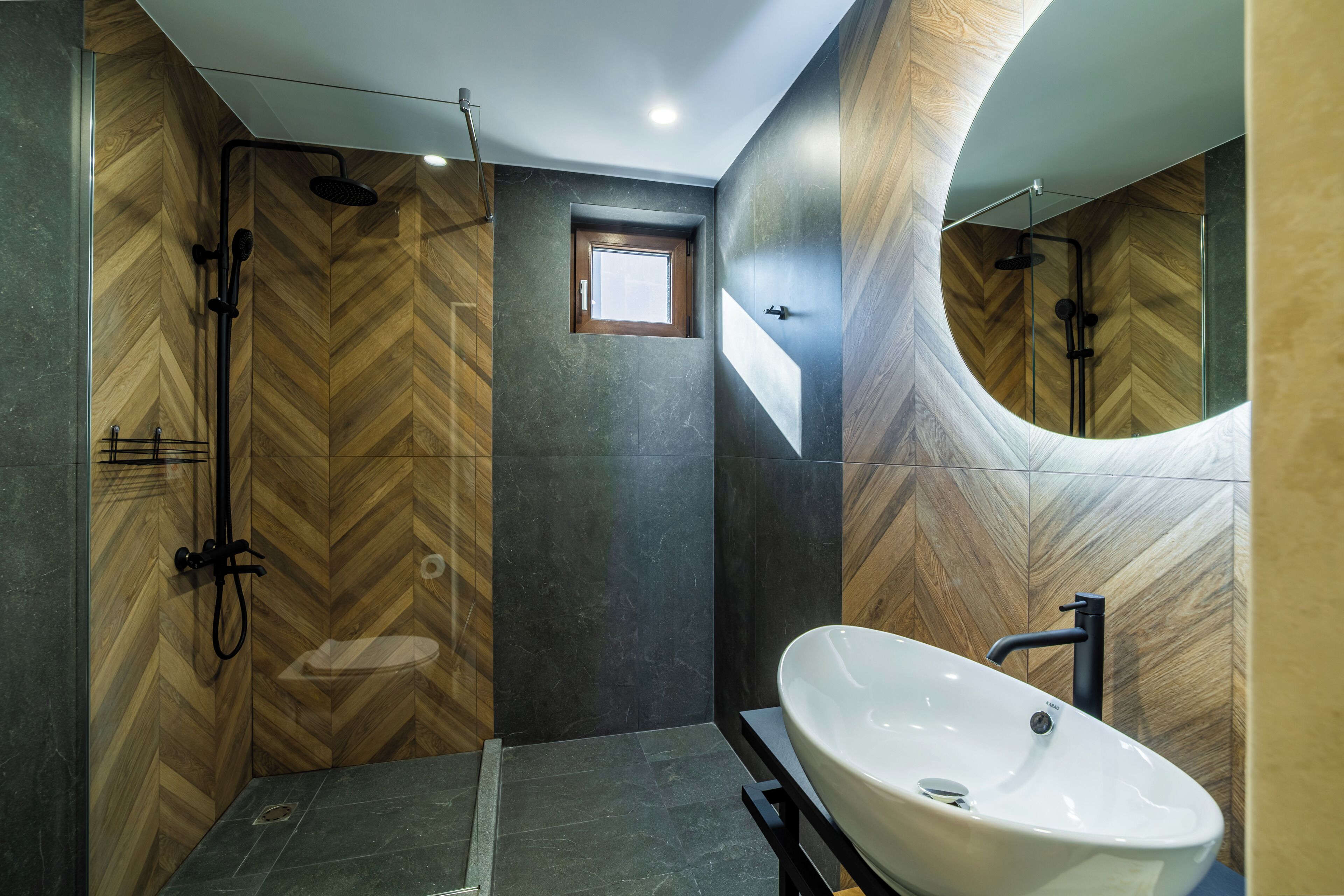 A modern bathroom with a herringbone wood pattern wall, a large white sink, and a shower area with a curved mirror.